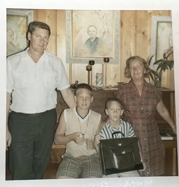 An old family photo. Four white people in front of a piano. Two blonde boys sit on a piano bench. The boy on the right holds a small bird and is smiling at the camera. The boy next to him has his mouth open. A white man in a short-sleeved button up shirt and black pants stands to the left, with a hand on the older boy’s back. A short, white blonde woman faces the camera and smiles. She is wearing a short-sleeved plaid red dress. Portraits of three boys flank the piano.