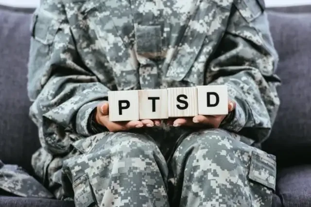 a zoom in of a person in military fatigues holding wooden blocks that spell out PTSD