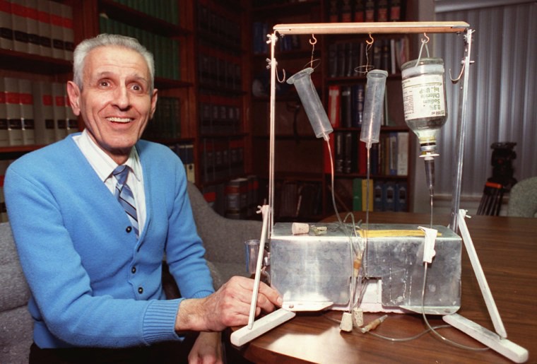 A picture courtesy NBC News of Dr. Jack Kevorkian, nicknamed Dr. Death, and his "suicide machine." A white man smiles looking upwards. He appears quite joyous, and he wears a pale blue sweater over a button up white shirt and crisp striped tie. His hair is gray and short. His hand is on his "machine" on a table next to him, which appears to have three vials hanging from a scaffolding of some kind and down into a container beneath.