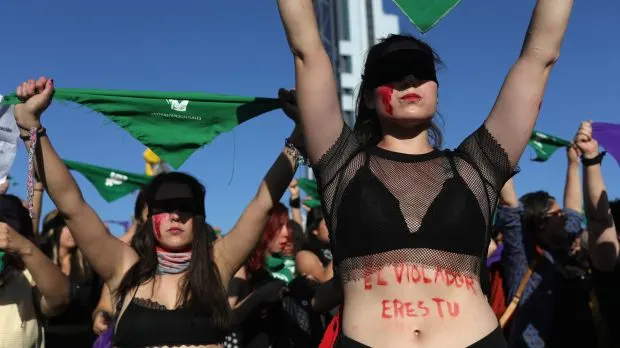 A photo of women in a group. The woman in foreground is wearing black bra top with fishnet over it. across her stomach in red is written: el violador eres tu. She has a black bandana covering her eyes, and red smeared on her face.