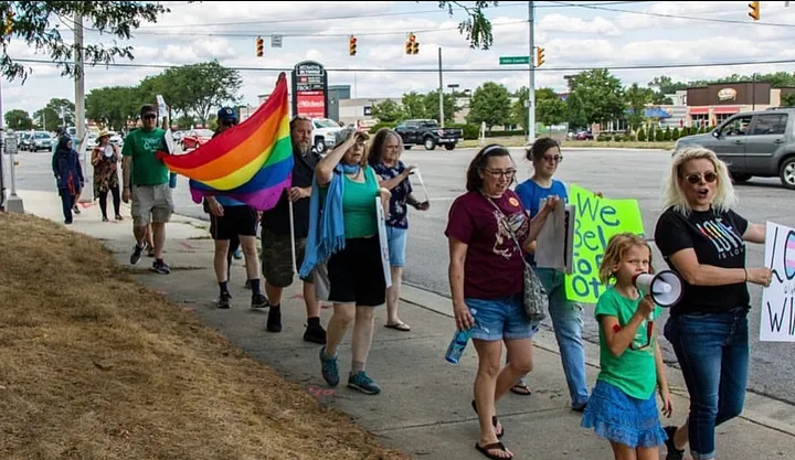 a photo of people walking down a road holding signs, a gay flag. a child and adult in front, child with megaphone, in foreground.