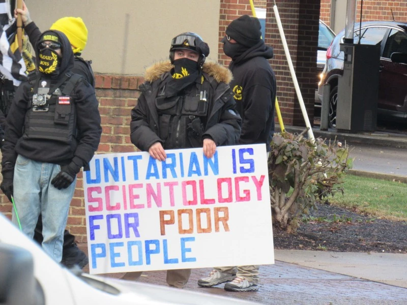 a bored looking white man in a thick black coat with most of his face covered holds a sign that reads: UNITARIAN IS SCIENTOLOGY FOR POOR PEOPLE