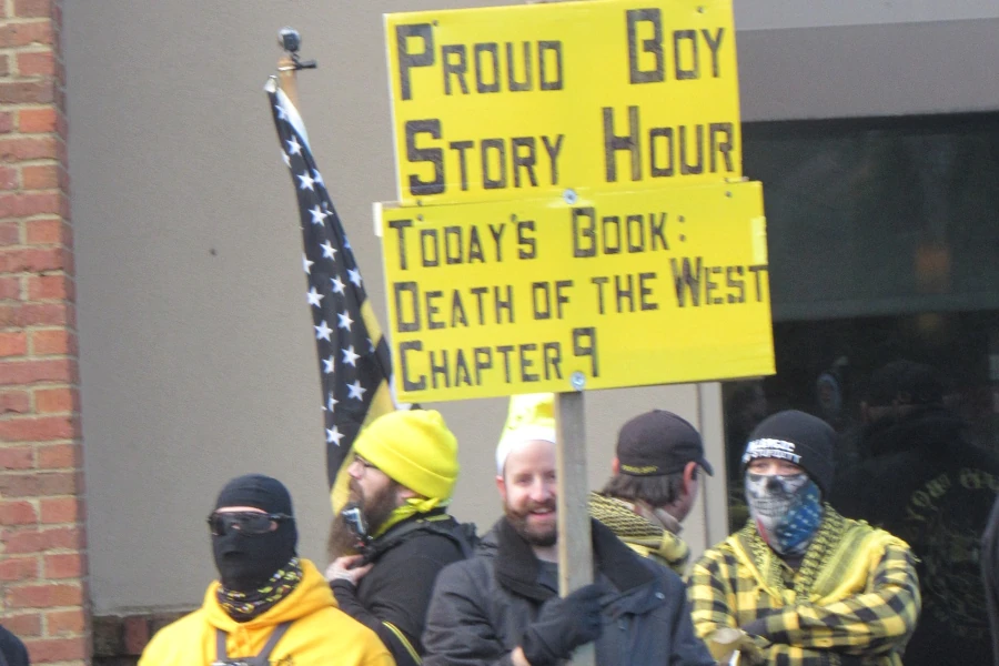 five white men. one with a full beard and mustache grins while holding a yellow sign in black letters reading: proud boy story hour today's book: death of the west chapter 9. other white men have faces covered. all wear yellow and black.