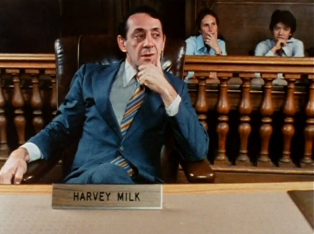 Image of white man in a blue suit and striped tie. He appears to be seated in a courtroom or public office. He looks intently on, chin in hands, bemused. 