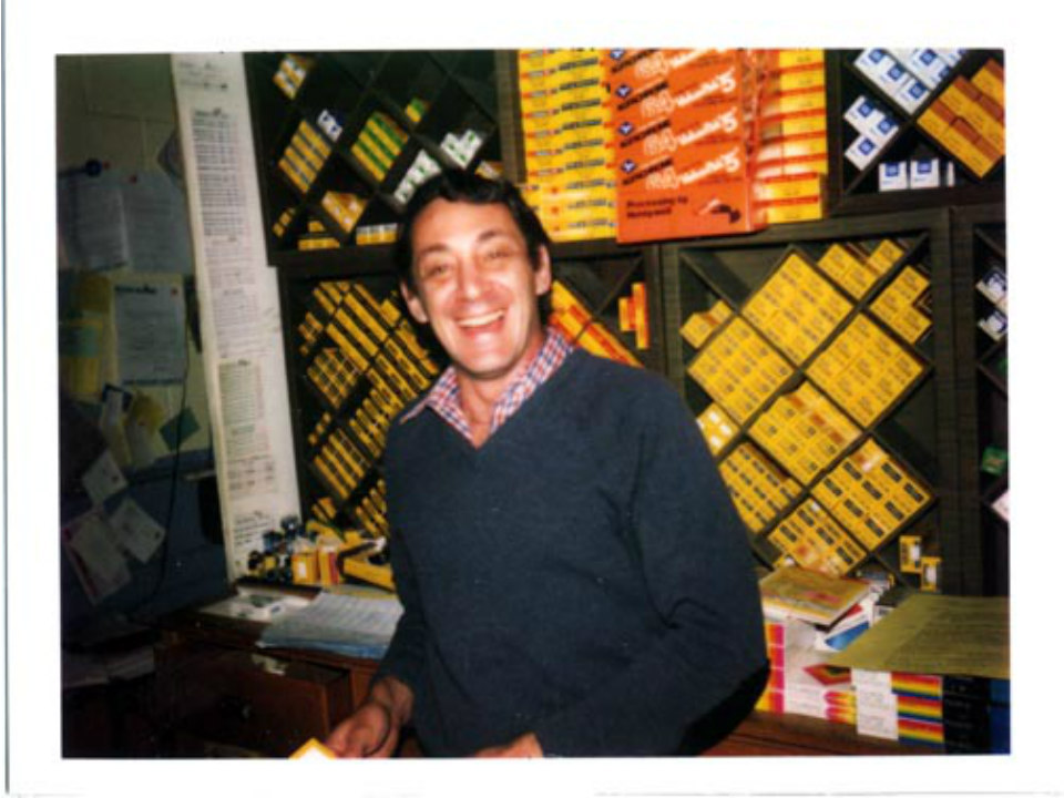 A color photo, an older one, somewhat blurry of a white man smiling broadly. He's wearing a navy blue, long sleeved sweater with button down underneath. He's got dark receding hairling and infectious grin. Behind him are shelves of camera film.