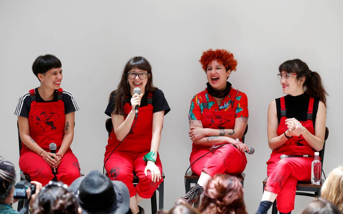 Image of four feminine figures dressed in black and red overalls laughing on a stage.