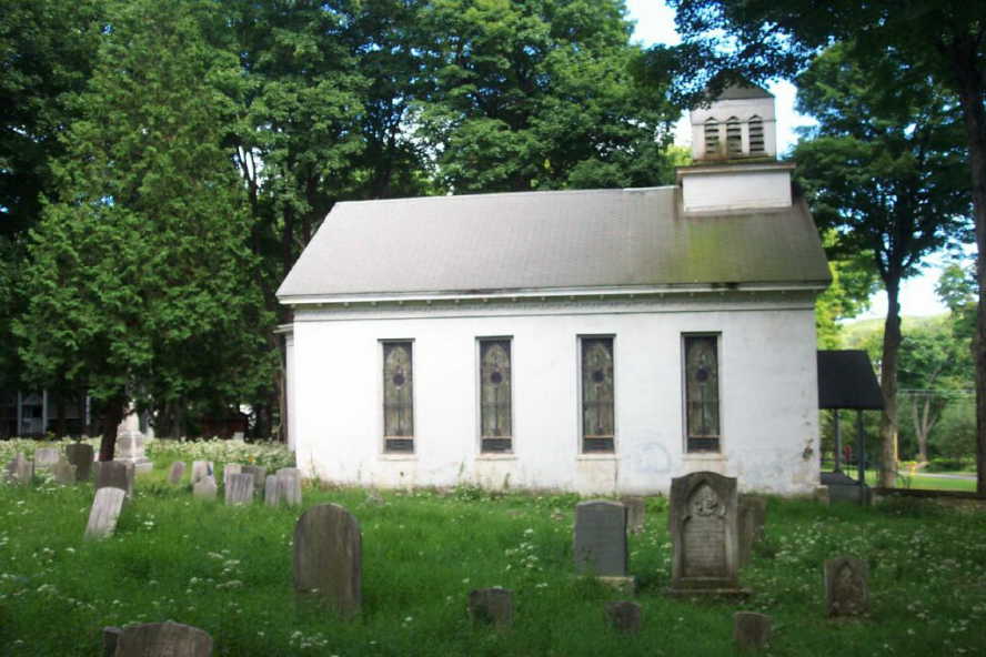 Image of old cemetery in the foreground and white church in distance with stained glass windows. The church is white and there is overgrowth and green trees shading it. 