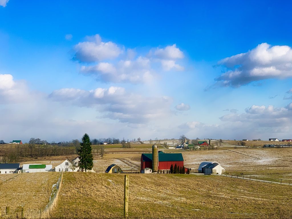 Winter farmland. top of image is blue sky and white clouds, with dead fields in the foreground. A barn and farmhouse in the background