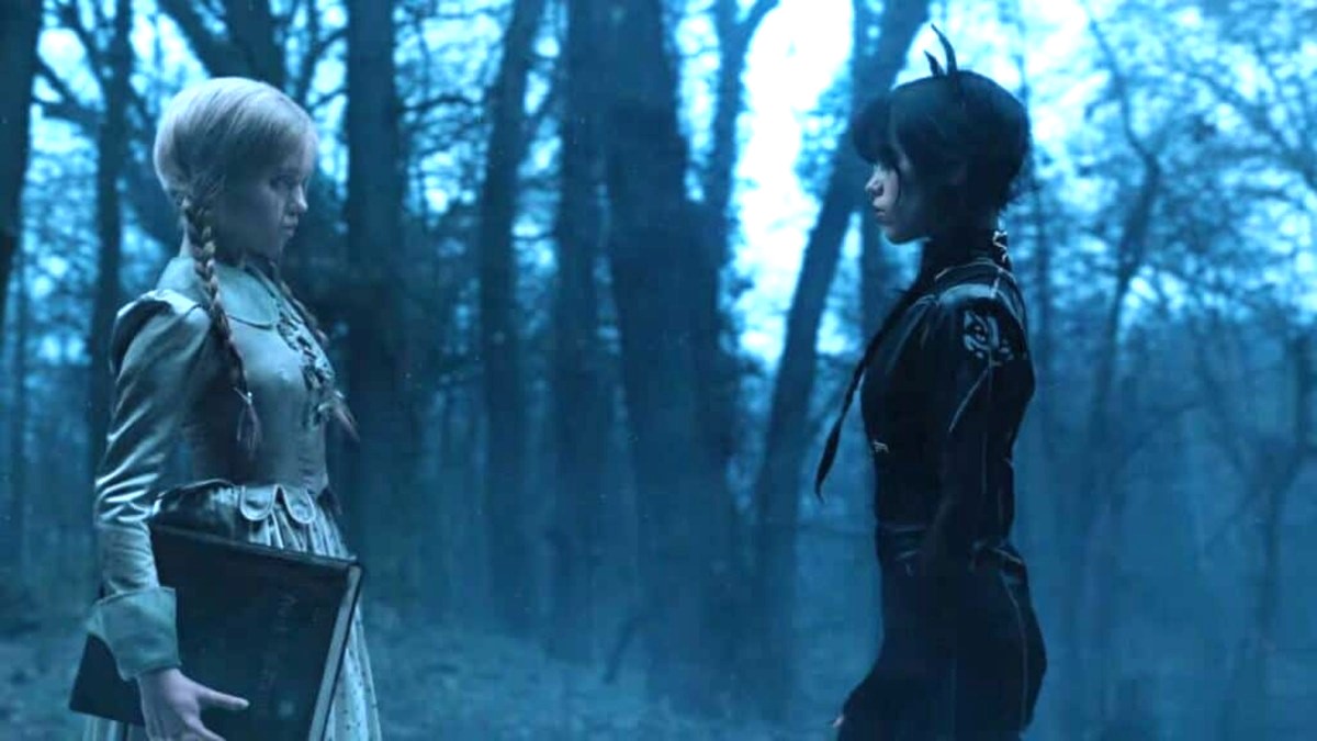 Image shows two girls standing facing one another. One with blonde braids and one with darker hair in the same style. They appear to stand in a woods, wearing older fashioned, feminine clothing. The blonde girl is covered neck to wrist wearing a dress. The darker haired girl faces her wearing more modern black clothing. They face one another and seen in profile