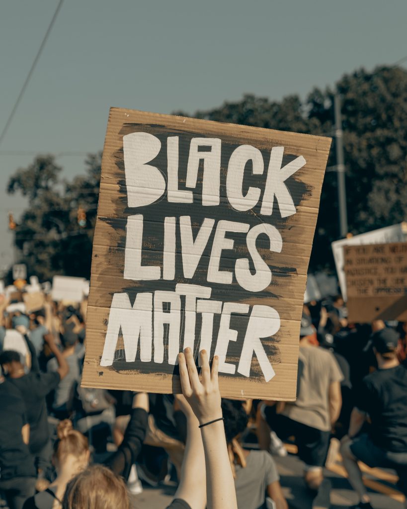 Image of a whilte hand holding a handmade sign reading in white print BLACK LIVES MATTER against black paint. Sign is brown cardboard