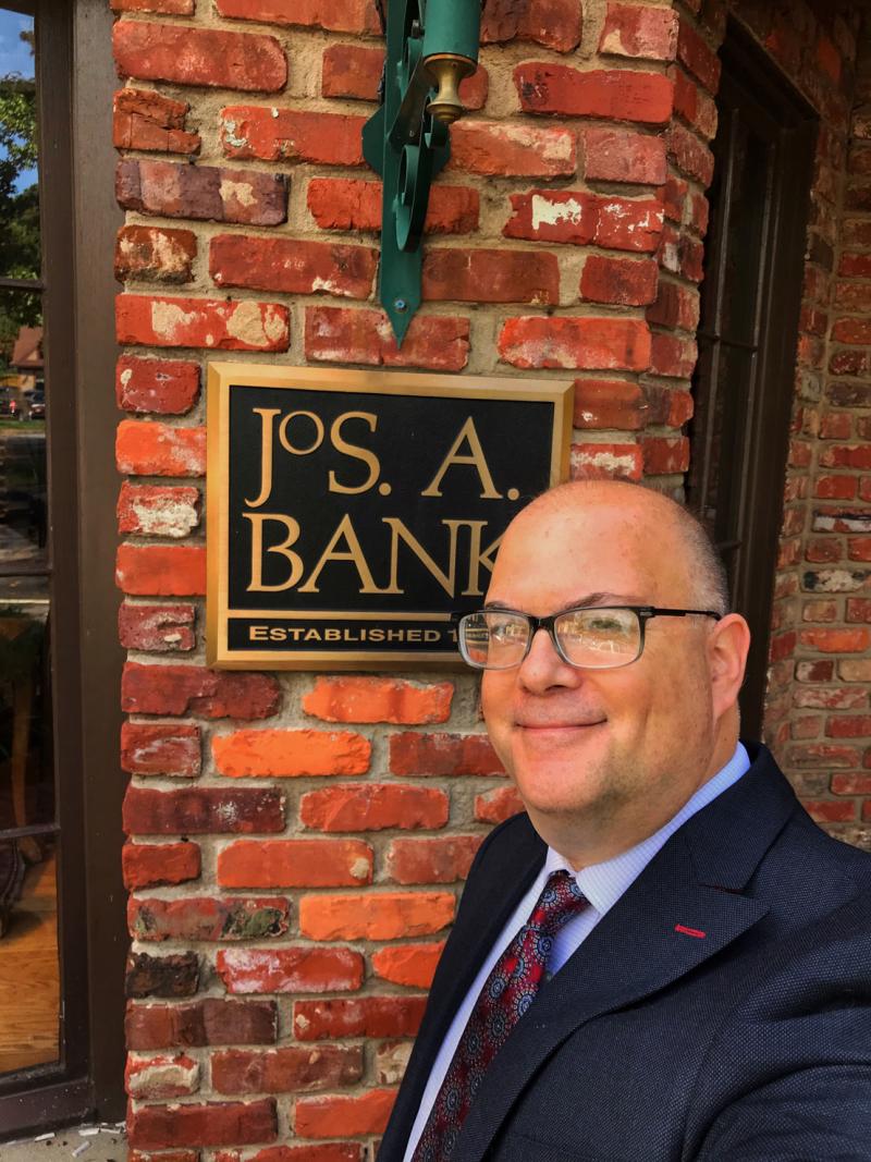 Image of a white man in a suite and tie taking selfie in front of Jos. A Bank. He's smiling, bald, wearing black rimmed glasses, middle aged