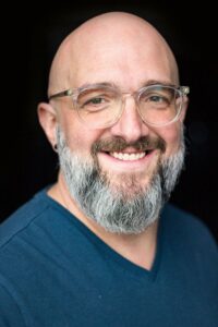 Image of smiling bald man with salt and pepper gray beard and clear glasses frame. From the Veritas website