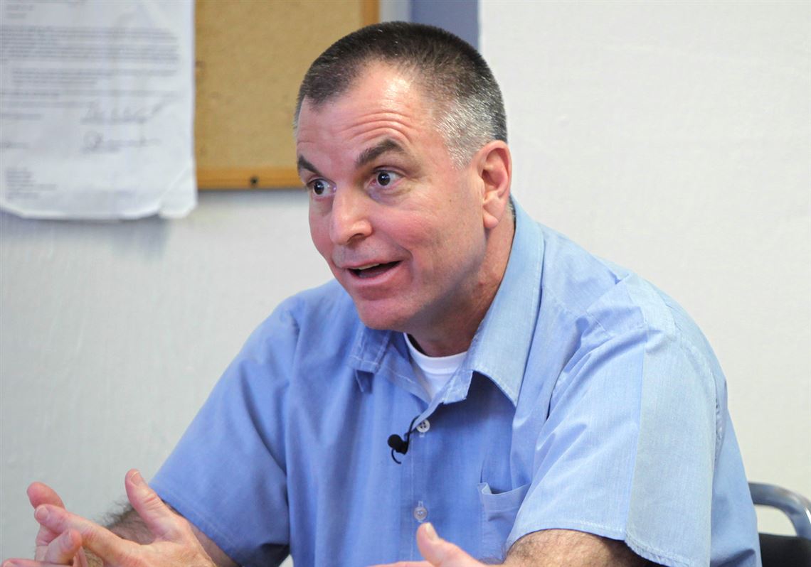 Image of middle-aged white man. He's facing to the left, appears to be in prison uniform of light blue collared shirt, arms crossed on table, mouth open, eyes wide. Image of Tom Noe in prison
