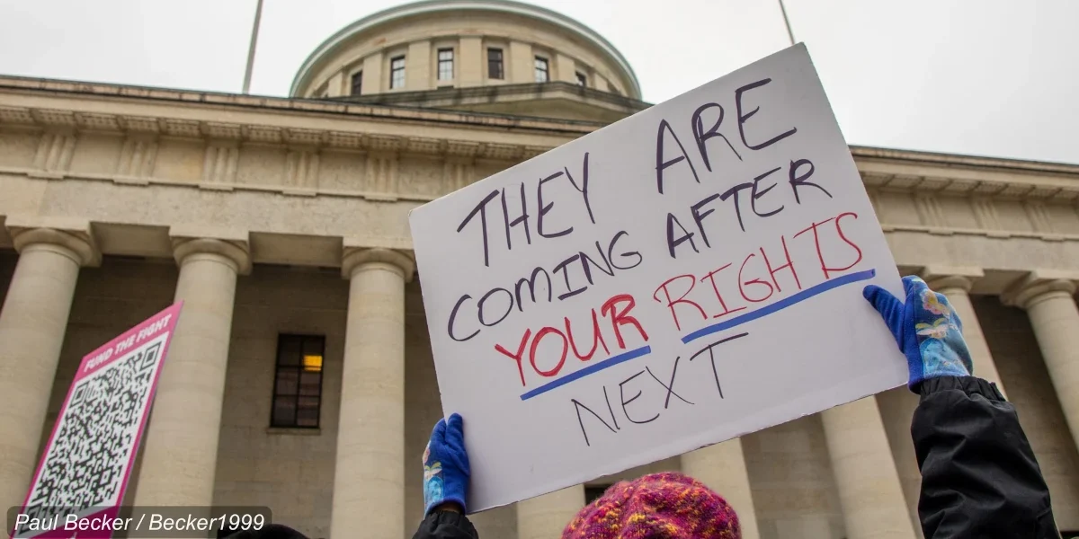 A protester's hands are visible holding a sign that reads they are coming for your rights next. The ohio statehouse with the ohio and american flag are in the background