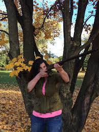 white woman with dark shoulder length hair, presumably Beth lear, holds shotgun aimed upwards with trees in background. From Beth Lear's LinkedIn