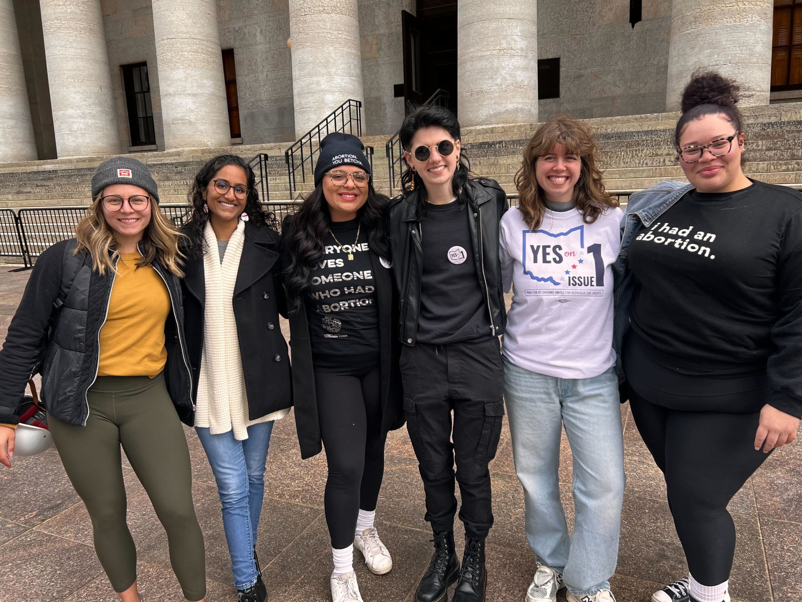 six individuals, arm-in-arm, in front of the Ohio Statehouse. One individual wears a t shirt that reads: someone you love has had an abortion. another visible t reads: I had an abortion. All are smiling and camera facing