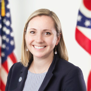 Image of smiling white woman with ohio and american flag in background. she's wearing a black blazer and has shoulder length straight dark blonde hair