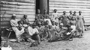 a black and white image of Black enslaved family in front of a home. Undated. About 20 people, men, women, and children, are photographed.