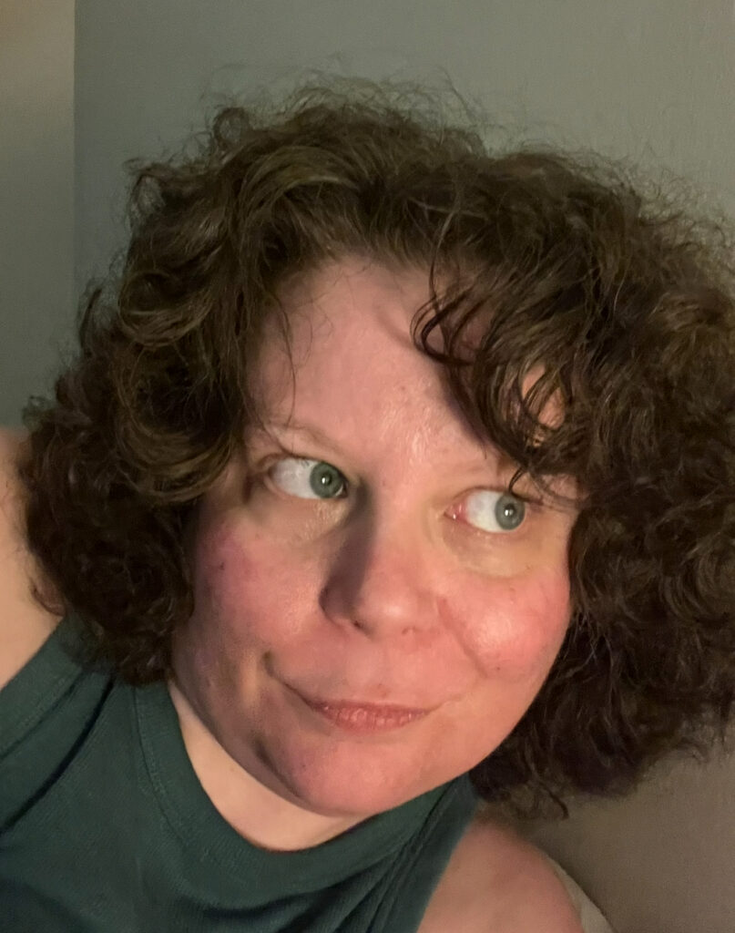 Image of L.J. Cunningham; close up of white person with short, dark, curly hair, smiling, and looking to the viewer's right, wearing dark green tank with top of shoulders visible.